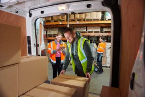 a male courier loads or unloads his van supervised by his female manager .. In the background a co-worker can be seen along the racking.
